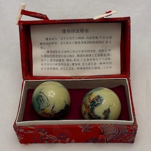 Pode incluir: Uma caixa vermelha retangular com interior branco contém duas bolas decorativas de exercício. As bolas são creme com cenas pintadas. Um pequeno cartão com texto chinês está dentro. A caixa tem um exterior vermelho estampado.