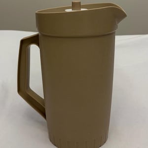 Op de afbeelding: Een beige kunststof kan met een verticaal handvat, een schenktuit en een deksel met een kleine centrale knop. De onderkant van de kan heeft een reeks verticale inkepingen rond de rand, wat een gestructureerd detail aan het ontwerp geeft.