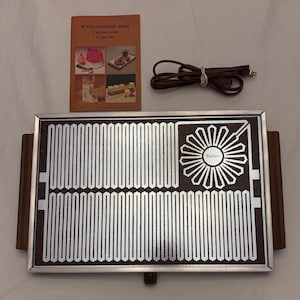 May include: A rectangular Salton warming tray with a dark brown surface featuring a silver metallic heating element pattern and a central flower-like design. It includes brown side handles, a detachable brown power cord, and an orange recipe booklet.