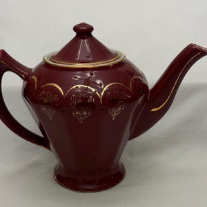 May include: A maroon ceramic teapot with a lid and a curved spout. The teapot has gold decorative accents and trim. The handle is curved and attached to the side. A classic design, the teapot is suitable for serving tea.