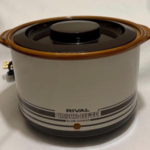 May include: A vintage Rival Crock-ette slow cooker. The appliance has a cream-colored body with brown and black accents, including a lid and a decorative band. The text "RIVAL CROCK-ETTE SLOW COOKER" is printed on the front.