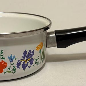 May include: A white enamel saucepan with a black handle and floral design. The pot features a silver rim and a pattern of colorful flowers, including red poppies, purple irises, and blue accents. The handle is ergonomically designed for easy grip.