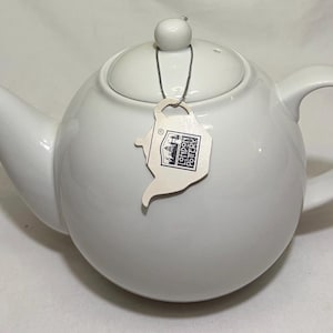 May include: A white ceramic teapot with a rounded body, curved spout, and a loop handle. The lid has a small knob. A tag with the words "London Pottery" hangs from the lid. The teapot is designed for brewing tea.