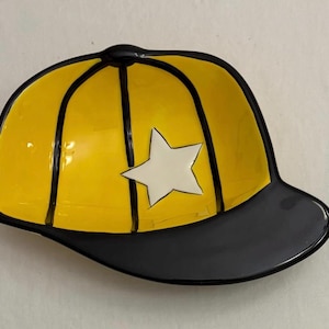 Puede incluir: Un plato decorativo de cerámica con forma de gorra de béisbol. La gorra es amarilla con ribetes negros y una estrella blanca. La visera es negra. El plato está diseñado para guardar objetos pequeños.