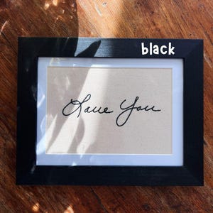 May include: Black framed artwork with the embroidered words "Love You" in cursive script. The artwork has a beige background and a white border. The frame is rectangular and the word "black" is written on the frame.