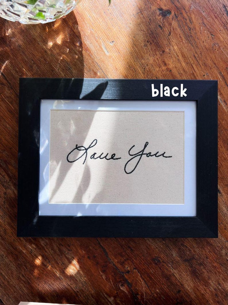 May include: Black framed artwork with the embroidered words "Love You" in cursive script. The artwork has a beige background and a white border. The frame is rectangular and the word "black" is written on the frame.