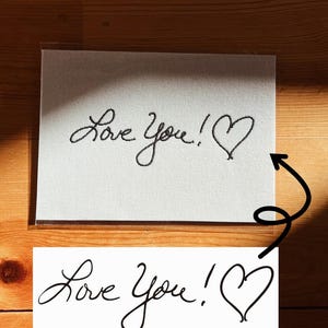 May include: A white card with the handwritten phrase "Love You!" and a heart symbol in black ink. The card is displayed on a wooden surface, with a second, similar card below.