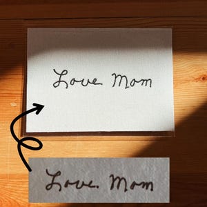 May include: A white canvas with the handwritten phrase "Love Mom" in black script. The canvas is in a clear plastic sleeve, and the image is on a wooden surface.