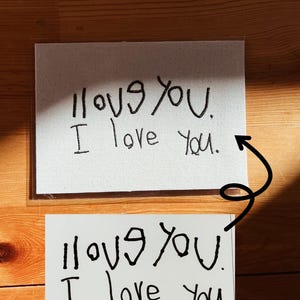 May include: Two white cards with the handwritten phrase "I love you." The top card is on a canvas-like material, while the bottom card is on plain white paper. Both cards are in black ink and are displayed on a wooden surface.