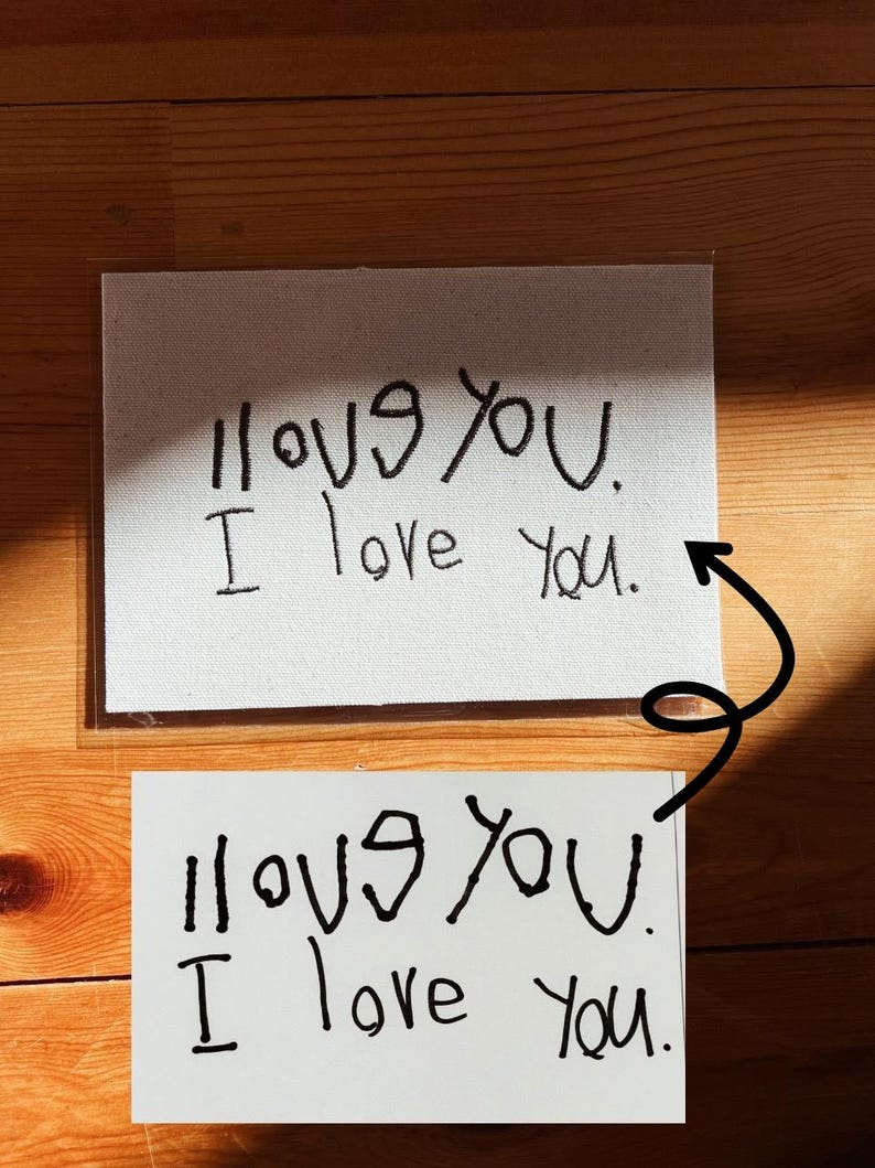 May include: Two white cards with the handwritten phrase "I love you." The top card is on a canvas-like material, while the bottom card is on plain white paper. Both cards are in black ink and are displayed on a wooden surface.