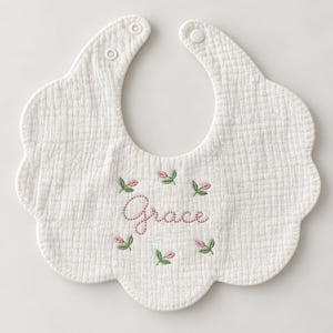 May include: A white scalloped-edge bib made of textured muslin fabric. The center features the embroidered name Grace in pink cursive script, surrounded by six small embroidered pink tulip buds with green stems and leaves. Two white snaps secure the bib.