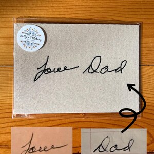 May include: A beige canvas with the embroidered words "Love Dad" in black cursive script. The canvas is in a clear plastic sleeve, with a round sticker in the upper left corner that reads "Kelly's Stitchery". Two smaller images of the handwriting are below.