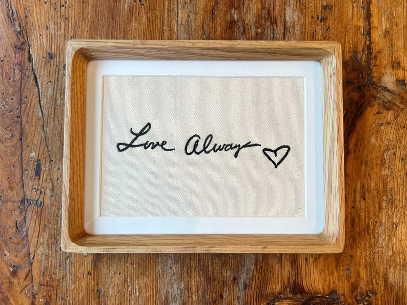 May include: A framed artwork with the words "Love Always" in black script, followed by a heart symbol. The artwork is set against a cream-colored fabric background, framed in a light-colored wooden frame.