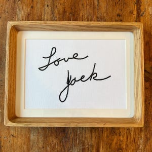 Könnte beinhalten: Ein gerahmtes Kunstwerk mit den Worten "Love Jack", die mit schwarzem Faden auf weißem Hintergrund gestickt sind. Der Rahmen besteht aus hellem Holz und das Stück wird auf einer Holzoberfläche präsentiert.