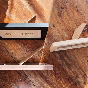 May include: Three small picture frames with easel backs are displayed on a wooden surface. One frame is black, and the other two are light wood. The black frame contains the handwritten text "Love You."