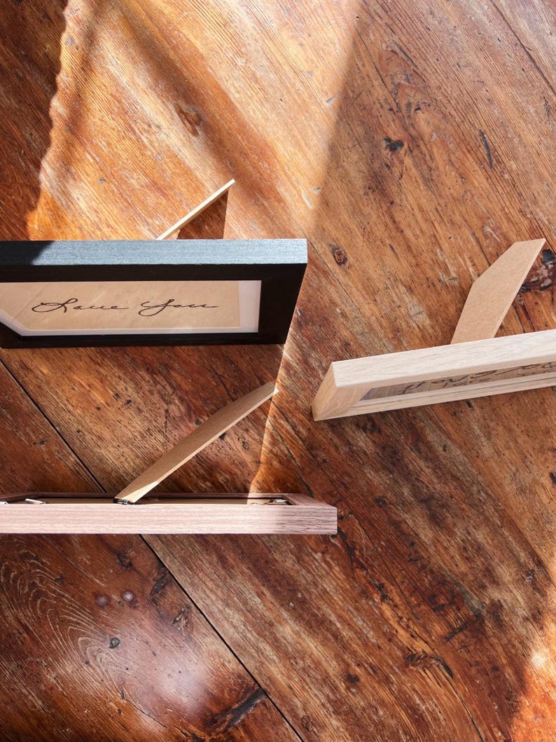 May include: Three small picture frames with easel backs are displayed on a wooden surface. One frame is black, and the other two are light wood. The black frame contains the handwritten text "Love You."
