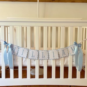 May include: A white crib with a banner that spells "WILLIAM" in blue letters. The banner is made of white scalloped-edge cards with blue lettering and is tied with blue ribbons. The crib is set against a wooden floor and wall.