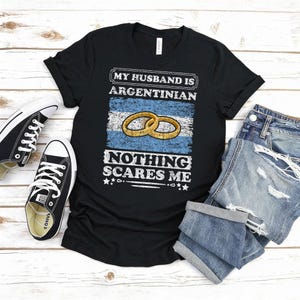 Puede incluir: Camiseta negra con el texto "MY HUSBAND IS ARGENTINIAN NOTHING SCARES ME" sobre un gr&aacute;fico de la bandera argentina y dos anillos de boda dorados. La camiseta se combina con zapatillas negras y pantalones cortos vaqueros azules.