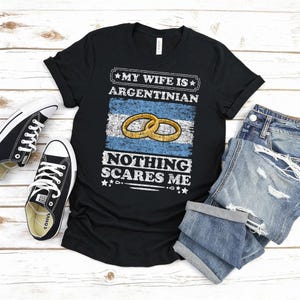 Puede incluir: Camiseta negra con el texto blanco "MY WIFE IS ARGENTINIAN NOTHING SCARES ME". El dise&ntilde;o incluye la bandera argentina y dos anillos de boda dorados. Un par de zapatillas Converse negras y unos pantalones cortos vaqueros doblados est&aacute;n junto a la camiseta.