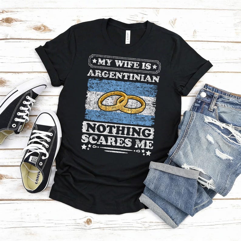 Puede incluir: Camiseta negra con el texto blanco "MY WIFE IS ARGENTINIAN NOTHING SCARES ME". El dise&ntilde;o incluye la bandera argentina y dos anillos de boda dorados. Un par de zapatillas Converse negras y unos pantalones cortos vaqueros doblados est&aacute;n junto a la camiseta.