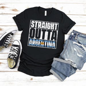 Puede incluir: Camiseta negra con el texto "STRAIGHT OUTTA ARGENTINA" en blanco, con un efecto desgastado. La bandera argentina se muestra debajo. La camiseta se combina con zapatillas negras y pantalones cortos vaqueros.