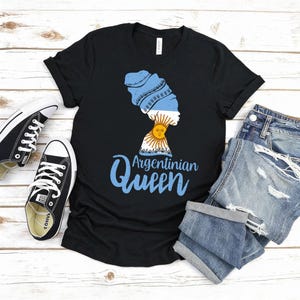 Puede incluir: Camiseta negra con un gr&aacute;fico de la silueta de una mujer con un pa&ntilde;uelo en la cabeza con los colores de la bandera argentina, un dise&ntilde;o de sol y el texto "Argentinian Queen" en azul. La camiseta se combina con pantalones cortos vaqueros y zapatillas negras.