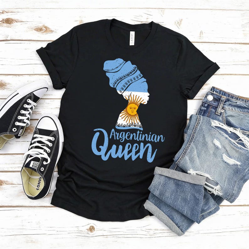 Puede incluir: Camiseta negra con un gr&aacute;fico de la silueta de una mujer con un pa&ntilde;uelo en la cabeza con los colores de la bandera argentina, un dise&ntilde;o de sol y el texto "Argentinian Queen" en azul. La camiseta se combina con pantalones cortos vaqueros y zapatillas negras.
