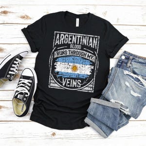 Puede incluir: Camiseta negra con el texto "ARGENTINIAN BLOOD RUNS THROUGH MY VEINS" y el dise&ntilde;o de la bandera argentina. La imagen tambi&eacute;n muestra zapatillas negras y pantalones cortos vaqueros.