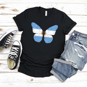 Puede incluir: Camiseta negra con un dise&ntilde;o de mariposa en los colores de la bandera argentina: azul claro y blanco. La imagen tambi&eacute;n muestra zapatillas negras y pantalones cortos de mezclilla, todo sobre una superficie de madera blanca.