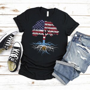 Puede incluir: Camiseta negra con un dise&ntilde;o de &aacute;rbol. La copa del &aacute;rbol presenta la bandera estadounidense, y las ra&iacute;ces muestran una bandera azul claro y blanca. La imagen incluye zapatillas negras y pantalones cortos de mezclilla sobre una superficie de madera blanca.