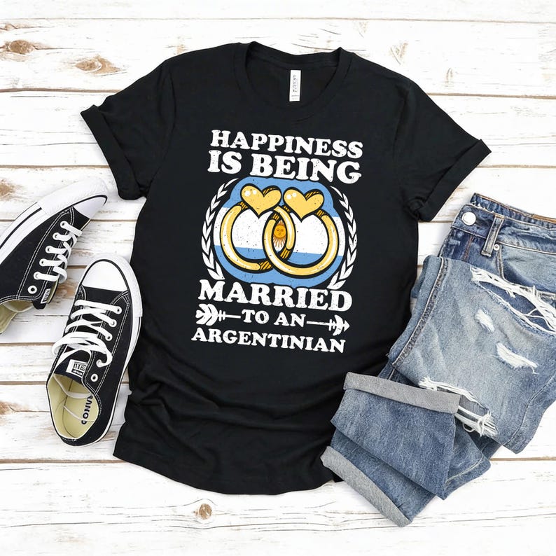 Puede incluir: Camiseta negra con el texto "HAPPINESS IS BEING MARRIED TO AN ARGENTINIAN". El dise&ntilde;o incluye dos anillos de boda con los colores de la bandera argentina y un coraz&oacute;n. Un par de zapatillas negras y unos pantalones cortos vaqueros azules est&aacute;n junto a la camiseta.