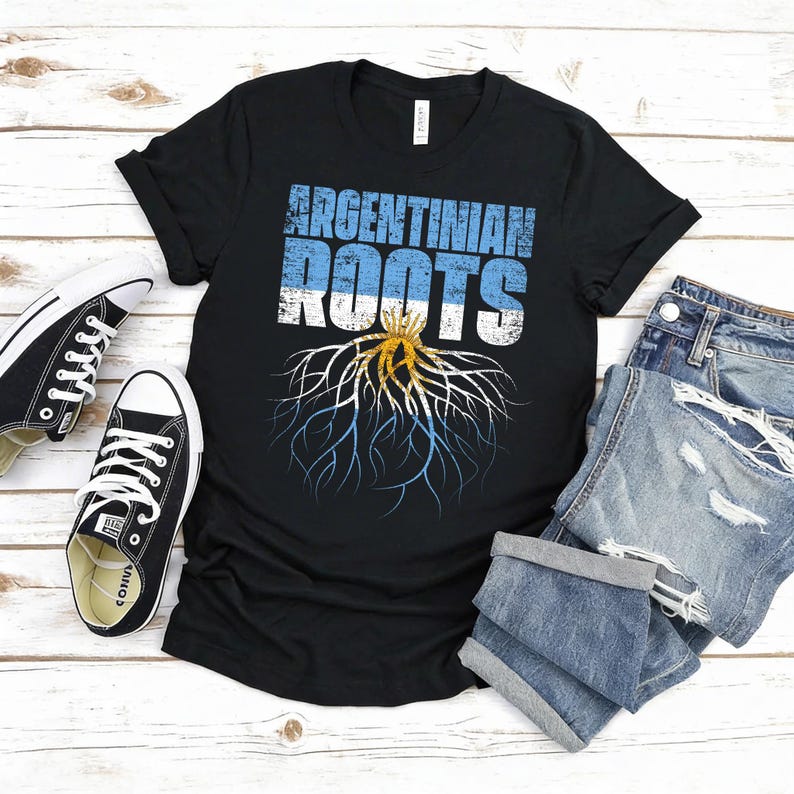 Puede incluir: Camiseta negra con el texto "ARGENTINIAN ROOTS" en azul y blanco, con un gr&aacute;fico de ra&iacute;ces con los colores de la bandera argentina. La camiseta se muestra con zapatillas negras y pantalones cortos vaqueros.