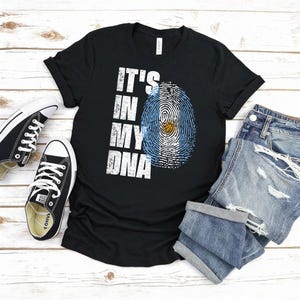 Puede incluir: Camiseta negra con el texto blanco "IT'S IN MY DNA", que presenta un dise&ntilde;o de huella dactilar con los colores de la bandera argentina. La camiseta se combina con zapatillas negras y pantalones cortos vaqueros, todo sobre una superficie de madera clara.