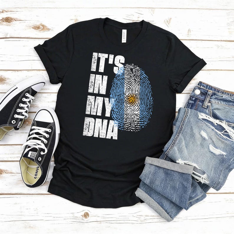 Puede incluir: Camiseta negra con el texto blanco "IT'S IN MY DNA", que presenta un dise&ntilde;o de huella dactilar con los colores de la bandera argentina. La camiseta se combina con zapatillas negras y pantalones cortos vaqueros, todo sobre una superficie de madera clara.