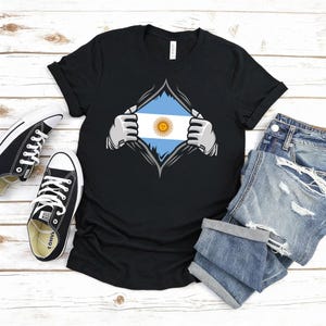 Puede incluir: Camiseta negra con un dise&ntilde;o gr&aacute;fico de la bandera argentina, como si se revelara desde debajo de la camiseta. El dise&ntilde;o incluye las franjas azules y blancas de la bandera y un sol amarillo. La imagen incluye zapatillas negras y pantalones cortos vaqueros.