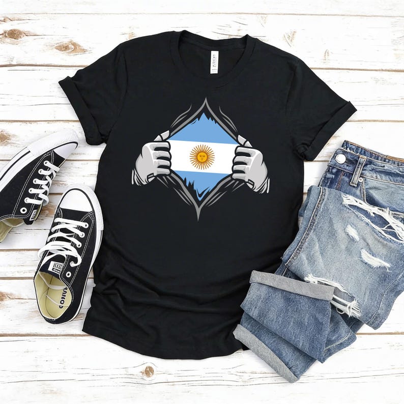Puede incluir: Camiseta negra con un dise&ntilde;o gr&aacute;fico de la bandera argentina, como si se revelara desde debajo de la camiseta. El dise&ntilde;o incluye las franjas azules y blancas de la bandera y un sol amarillo. La imagen incluye zapatillas negras y pantalones cortos vaqueros.