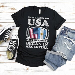 Puede incluir: Camiseta negra con el texto "I MAY LIVE IN THE USA BUT MY STORY BEGAN IN ARGENTINA." El dise&ntilde;o presenta las banderas estadounidense y argentina. Un par de zapatillas negras y unos vaqueros azules doblados tambi&eacute;n est&aacute;n en la imagen.