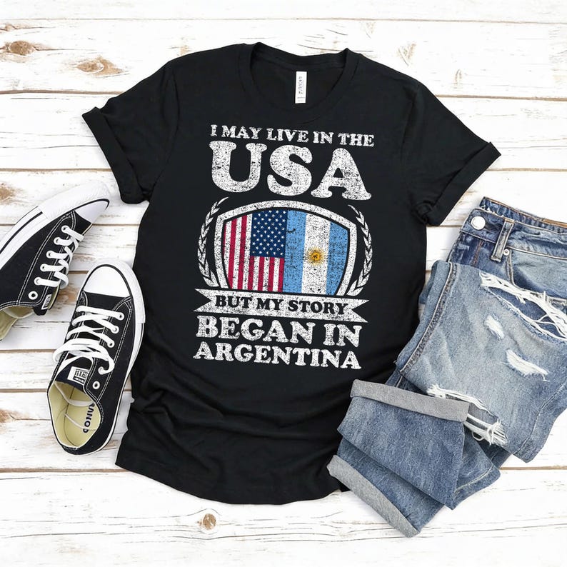 Puede incluir: Camiseta negra con el texto "I MAY LIVE IN THE USA BUT MY STORY BEGAN IN ARGENTINA." El dise&ntilde;o presenta las banderas estadounidense y argentina. Un par de zapatillas negras y unos vaqueros azules doblados tambi&eacute;n est&aacute;n en la imagen.