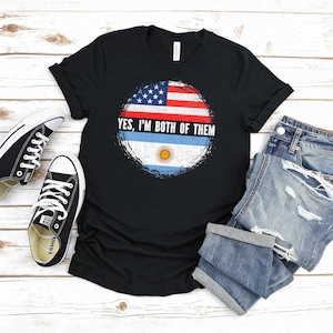 Puede incluir: Camiseta negra con un gr&aacute;fico circular desgastado con las banderas estadounidense y argentina, y el texto "YES, I'M BOTH OF THEM". La camiseta se combina con zapatillas negras y pantalones cortos vaqueros.