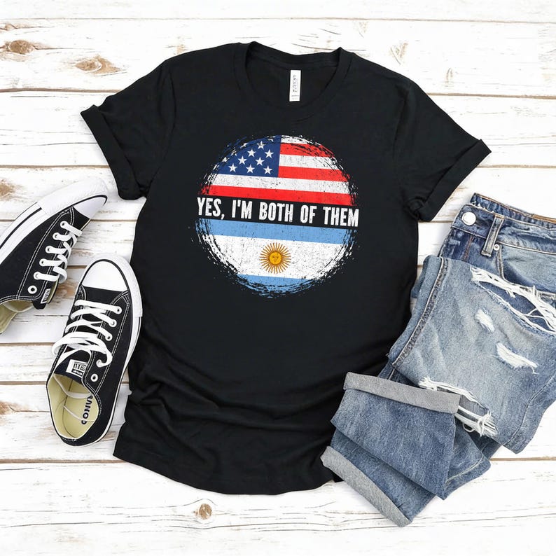 Puede incluir: Camiseta negra con un gr&aacute;fico circular desgastado con las banderas estadounidense y argentina, y el texto "YES, I'M BOTH OF THEM". La camiseta se combina con zapatillas negras y pantalones cortos vaqueros.
