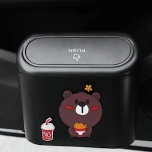 May include: Black car trash bin with a push-button lid. The bin features a cartoon bear holding fries and a drink graphic. The lid has the word "PUSH" and a hand symbol.