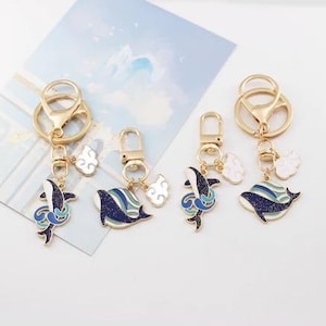 May include: Four gold-coloured keyrings with whale charms. Each charm features a blue and white whale design with a cloud accent. The keyrings have a lobster clasp and a split ring for attaching keys.