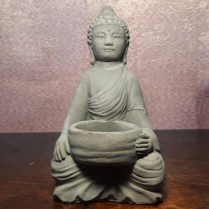 May include: A gray concrete Buddha statue sitting in a meditative pose, holding a small bowl in its lap. The statue has a serene expression and a traditional headdress.