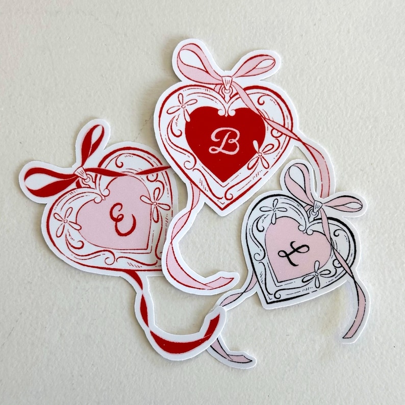 K&ouml;nnte beinhalten: Drei herzf&ouml;rmige Aufkleber mit dekorativen R&auml;ndern und B&auml;ndern. Jeder Aufkleber zeigt einen anderen Anfangsbuchstaben: "B", "E" und "F". Die Herzen sind in Rot-, Rosa- und Wei&szlig;t&ouml;nen gehalten, mit einer Schleife oben.