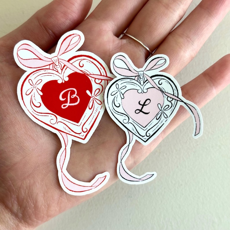 K&ouml;nnte beinhalten: Zwei herzf&ouml;rmige Aufkleber mit Schleifen- und Blumendesign. Ein Aufkleber ist rot mit wei&szlig;em Umriss und dem Buchstaben "B". Der andere ist rosa mit schwarzem Umriss und dem Buchstaben "L". Beide haben rosa B&auml;nder.