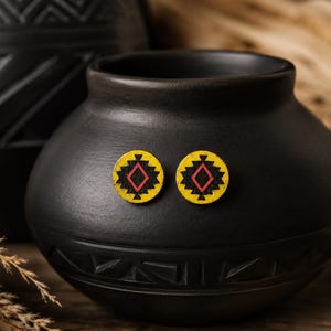 May include: A pair of circular stud earrings featuring a yellow background with a black geometric star pattern and a central red diamond outline. The earrings are displayed on the front of a matte black ceramic pot with a textured base.