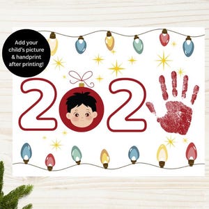 Pode incluir: Uma impressão com tema natalino com os números vermelhos 2023, o rosto de uma criança em uma bola vermelha e uma impressão de mão vermelha. A impressão inclui luzes de corda e o texto "Adicione a foto e a impressão da mão do seu filho após a impressão!"
