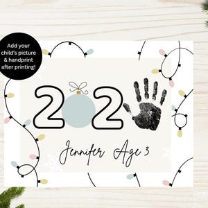 May include: A printable Christmas-themed design with the year 2020, a handprint, and the text "Jennifer Age 3." The design includes a light blue ornament and string lights. The text "Add your child's picture & handprint after printing!" is also included.