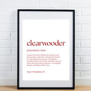 May include: A framed print with the word "clearwooder" in red, defined as a special time for Phillies fans migrating south. The definition includes details about baseball, salt air, and Wawa coffee. Origin: Philadelphia, PA.
