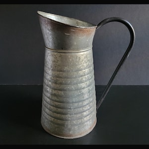 May include: A vintage metal pitcher with a black handle. The pitcher has a wide, angled spout and a ribbed body. The metal has a weathered, aged appearance with some rust around the rim. The handle is curved and attached to the side of the pitcher.